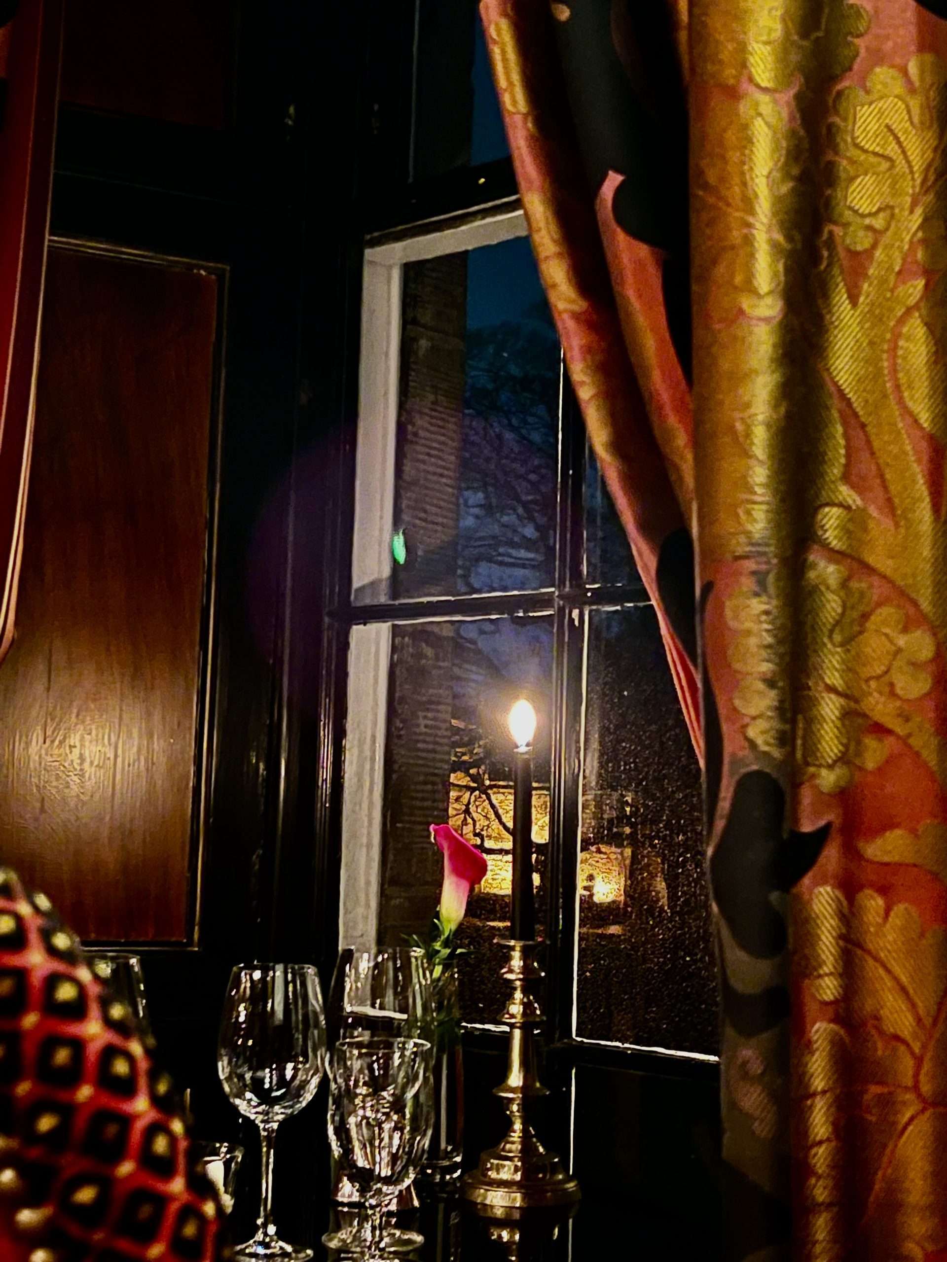 Tavolo nero con bicchieri, fiore in un vaso e candela accesa davanti a finestra con tendone rosso e giallo
