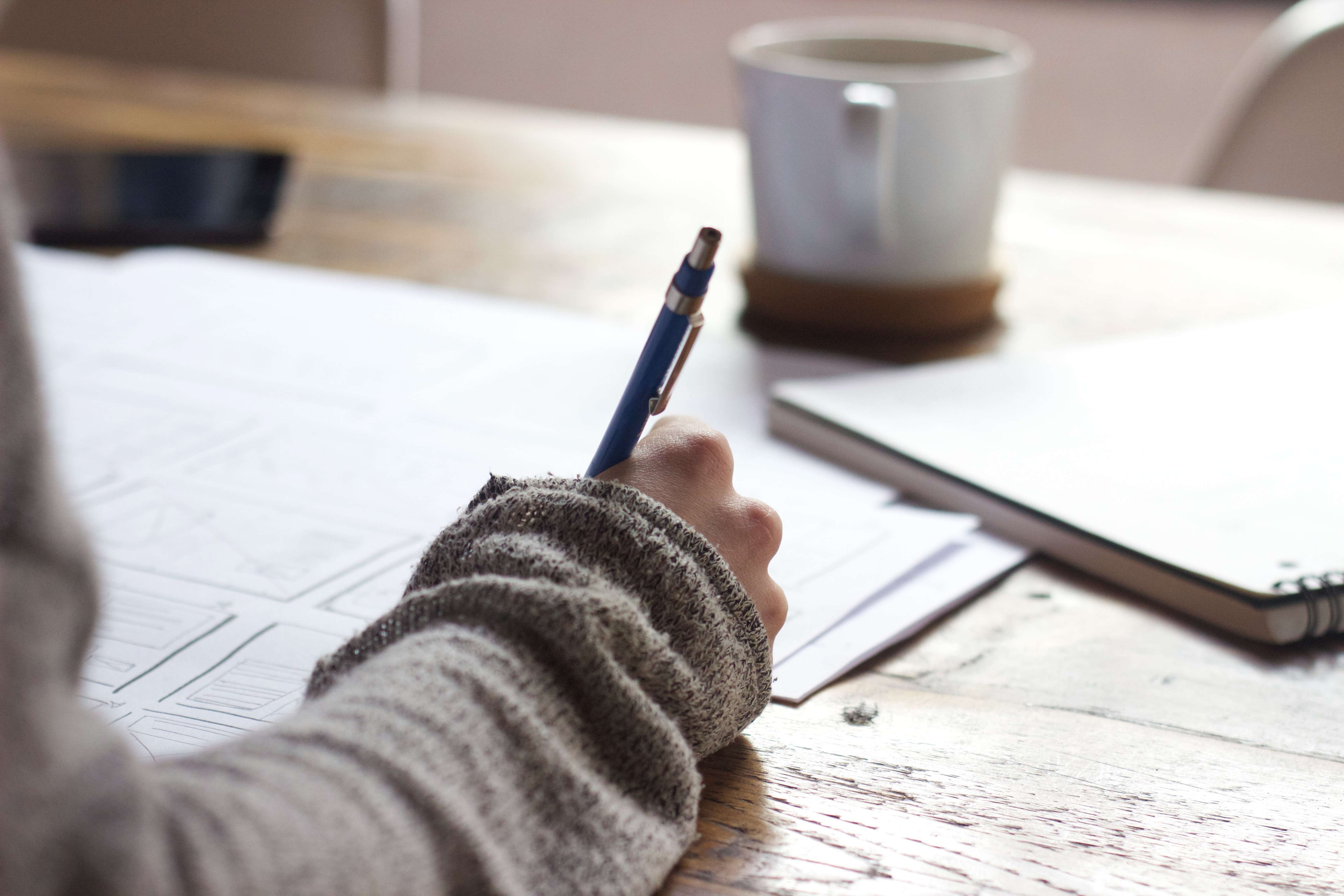 Persona che scrive su un foglio su un tavolo di legno con tazza di ceramica affianco per visto studenti UK