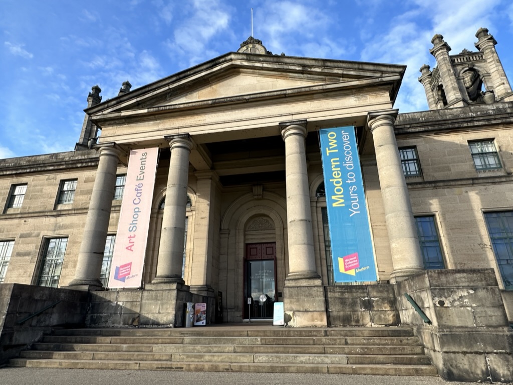Facciata d'ingresso in pietra arenaria della Scottish National Gallery of Modern Art, Modern Two, a Edimburgo, con colonne e striscioni colorati sotto un cielo blu con nuvole.