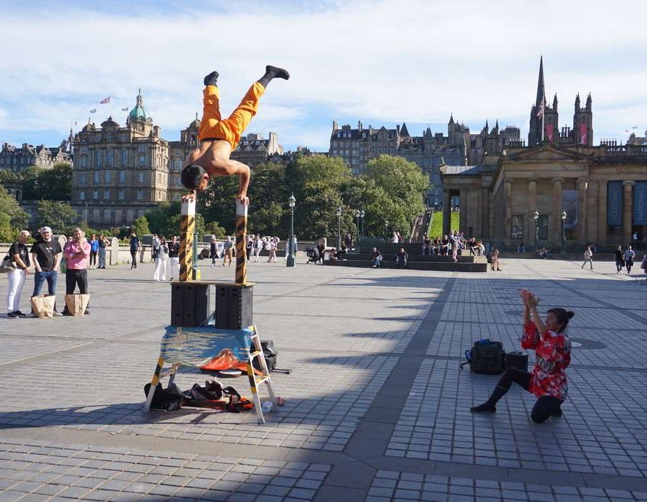 Strada a Edimburgo con artista a testa in giu e gente che osserva per festival di Edimburgo