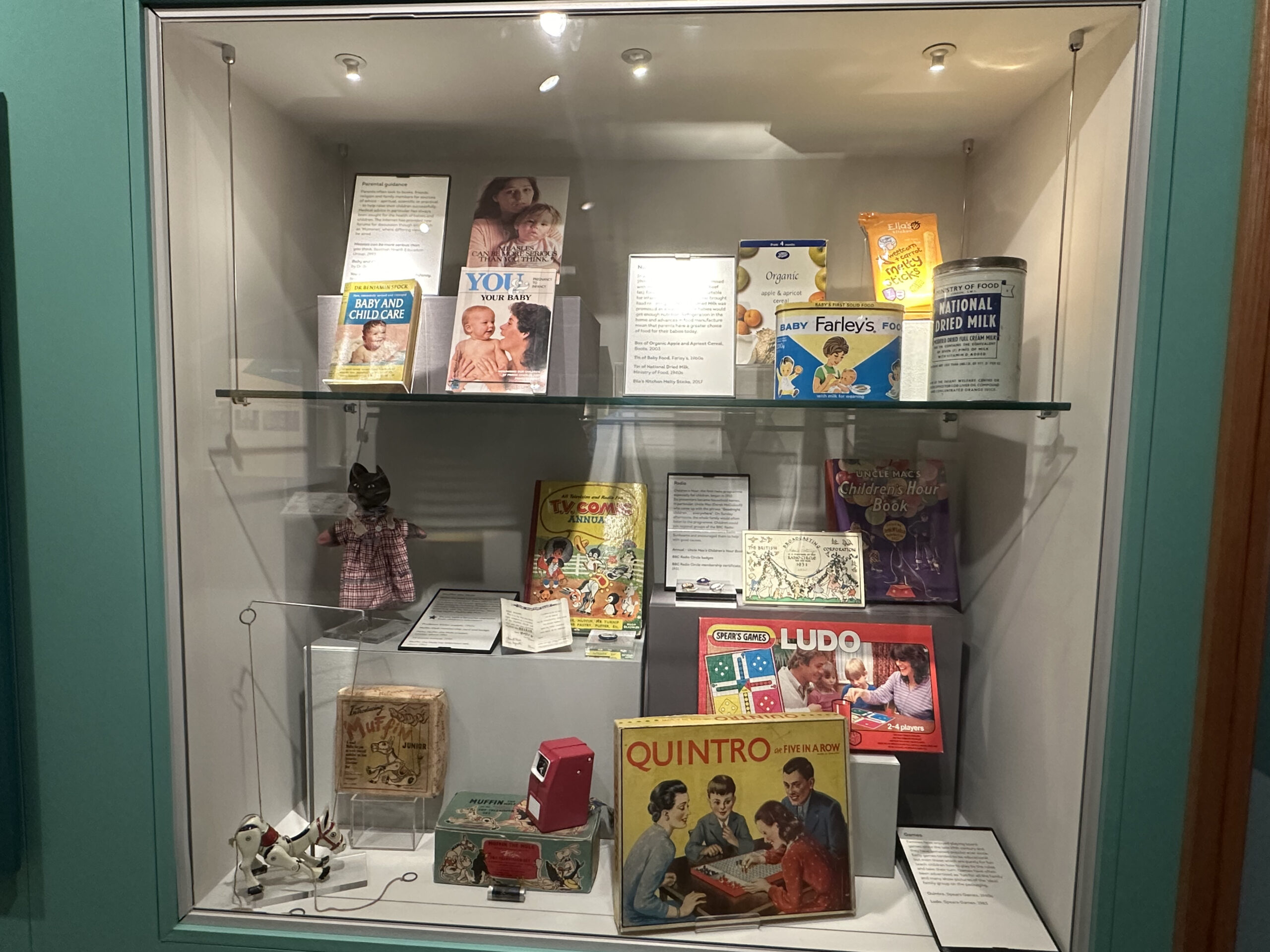 Una vetrina interna del Museo dell'Infanzia a Edimburgo, riempita con oggetti storici e nostalgici come latte di latte in polvere, libri per bambini e giochi da tavolo. L'immagine mostra l'atmosfera storica, con iscrizioni dorate leggibili come "NOSTALGIA & INFANZIA" e "UN TUFFO NEI RICORDI STORICI" integrate visivamente.
