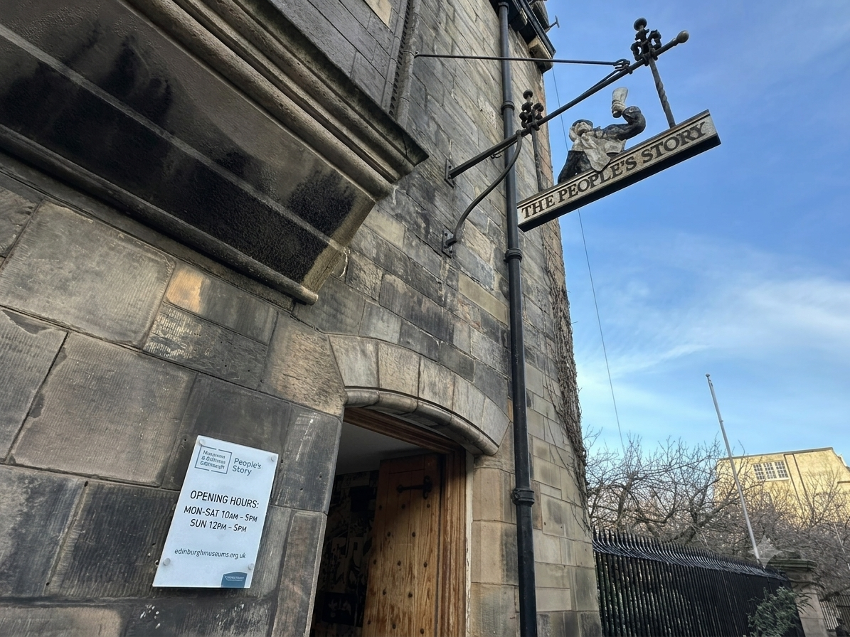 L'ingresso del People's Story Museum a Edimburgo, una veduta dell'architettura in pietra e del cartello informativo. L'immagine mostra l'atmosfera storica, con iscrizioni dorate leggibili come "THE PEOPLE'S STORY MUSEUM" e "EXPLORE EDINBURGH'S HISTORY" integrate visivamente.